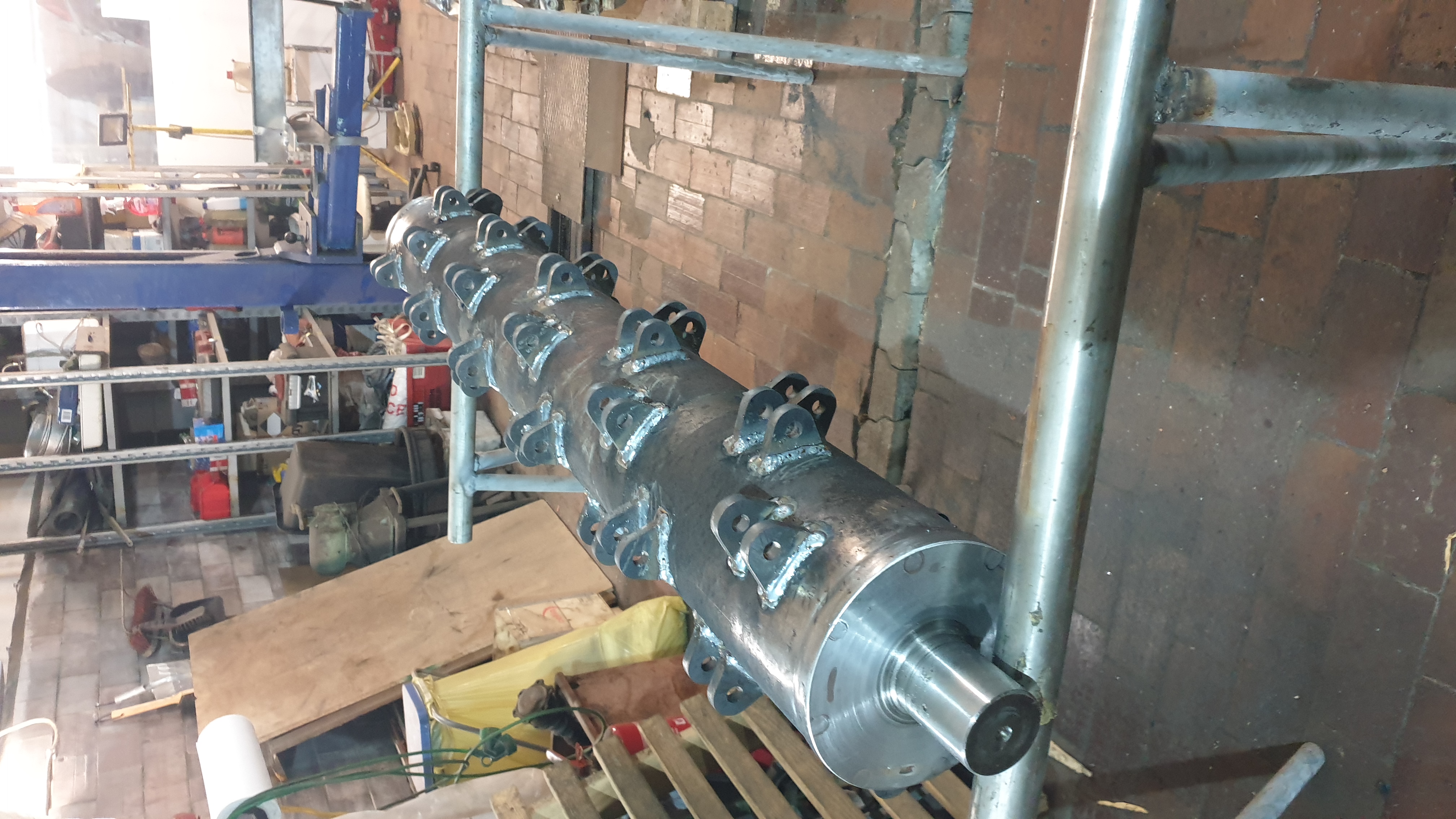 Metalowy walec z przyspawanymi uchwytami, umieszczony na rusztowaniu w warsztacie, widoczne ślady spawania i obróbki metalu.