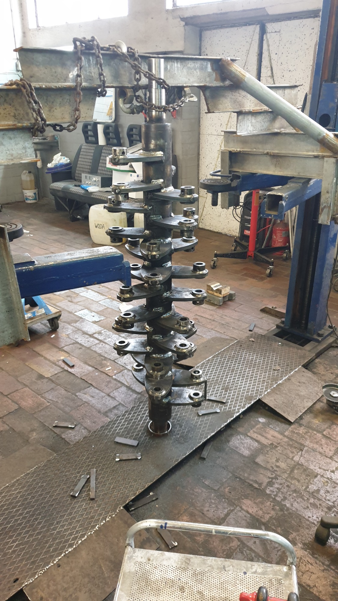 Surowy, metalowy element konstrukcyjny w trakcie spawania, umieszczony w warsztacie z widocznymi śladami obróbki i narzędziami, podwieszony na łańcuchach do suwnicy, z widocznymi spawami...