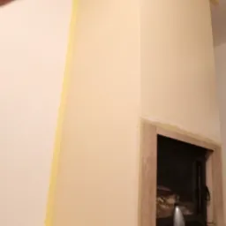 Obudowa kominka w trakcie malowania, zabezpieczona taśmą malarską, widoczne akcesoria malarskie i butelka wody.