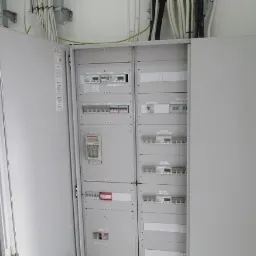 Otwarta szara szafa elektryczna z zainstalowanymi licznikami i bezpiecznikami, widoczne kable w białej ścianie.