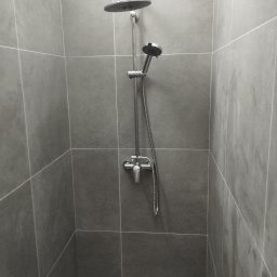 Katarzyna Grzesiek Fragles Usługi remontowo budowalne - Nowoczesna kabina prysznicowa wyłożona szarymi, kwadratowymi płytkami z widoczną armaturą: deszczownicą, słuchawką prysznicową i baterią.