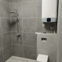 Katarzyna Grzesiek Fragles Usługi remontowo budowalne - Szara łazienka z kabiną prysznicową typu walk-in, podwieszaną toaletą i bojlerem elektrycznym, wykończona płytkami w jednolitym odcieniu szarości.