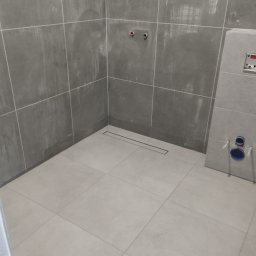 Katarzyna Grzesiek Fragles Usługi remontowo budowalne - Szara łazienka w trakcie remontu z widocznym odpływem liniowym w podłodze, surowymi przyłączami wodnymi na ścianie i zabudową geberit.