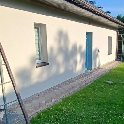 Katarzyna Grzesiek Fragles Usługi remontowo budowalne - Świeżo otynkowana elewacja budynku jednorodzinnego z niebieskimi drzwiami wejściowymi, widoczna drabina oparta o ścianę, trawnik z koniczyną w pierwszym planie.