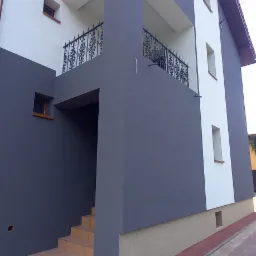 Dwukondygnacyjny dom z szarą i białą elewacją, balkon z ozdobną, metalową balustradą i schody wejściowe z beżowymi płytkami.