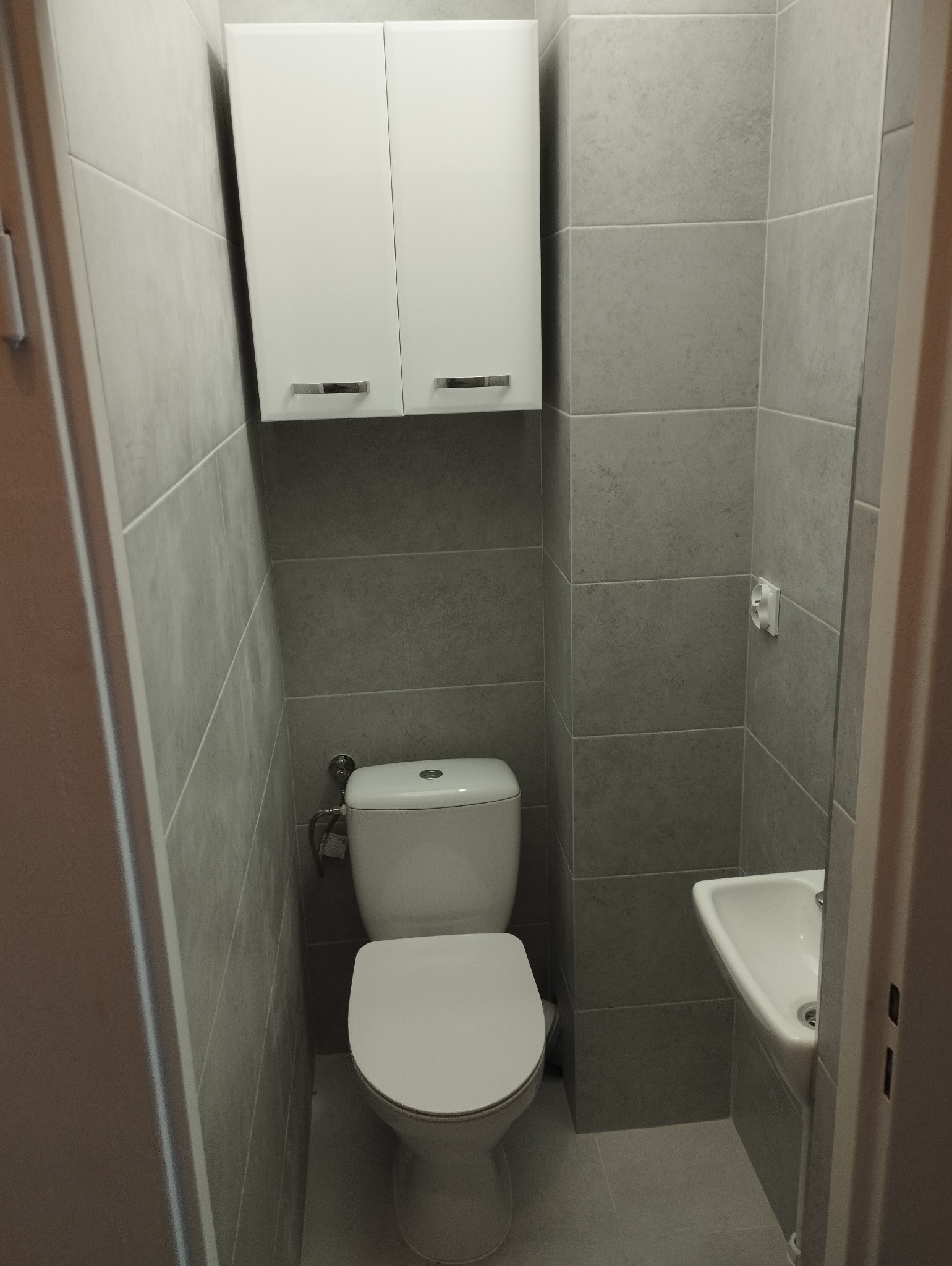 Nowoczesna, mała łazienka w szarościach z podwieszaną toaletą i szafką nad nią, oraz małą umywalką narożną.