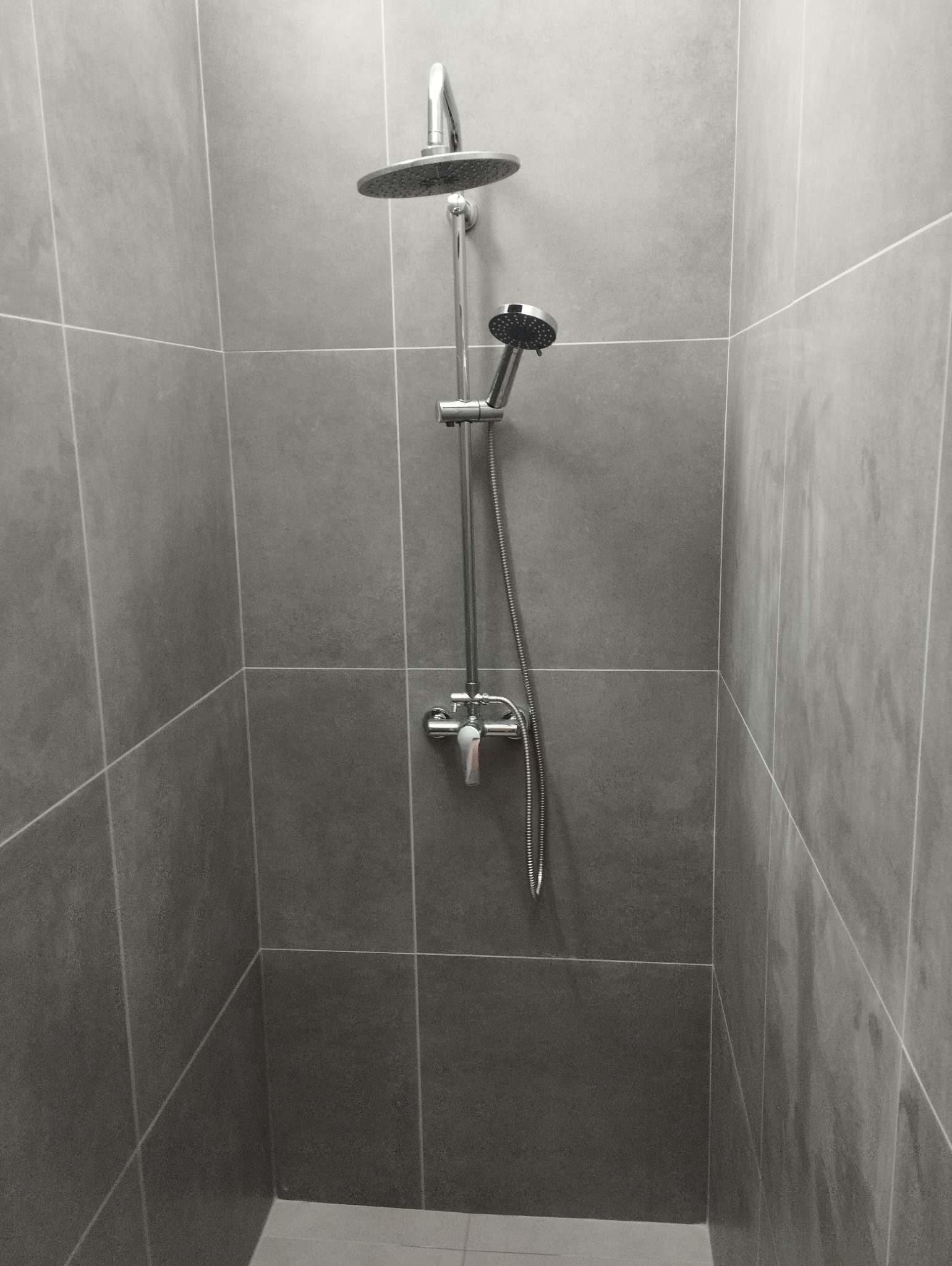 Nowoczesna kabina prysznicowa wyłożona szarymi, kwadratowymi płytkami z widoczną armaturą: deszczownicą, słuchawką prysznicową i baterią.