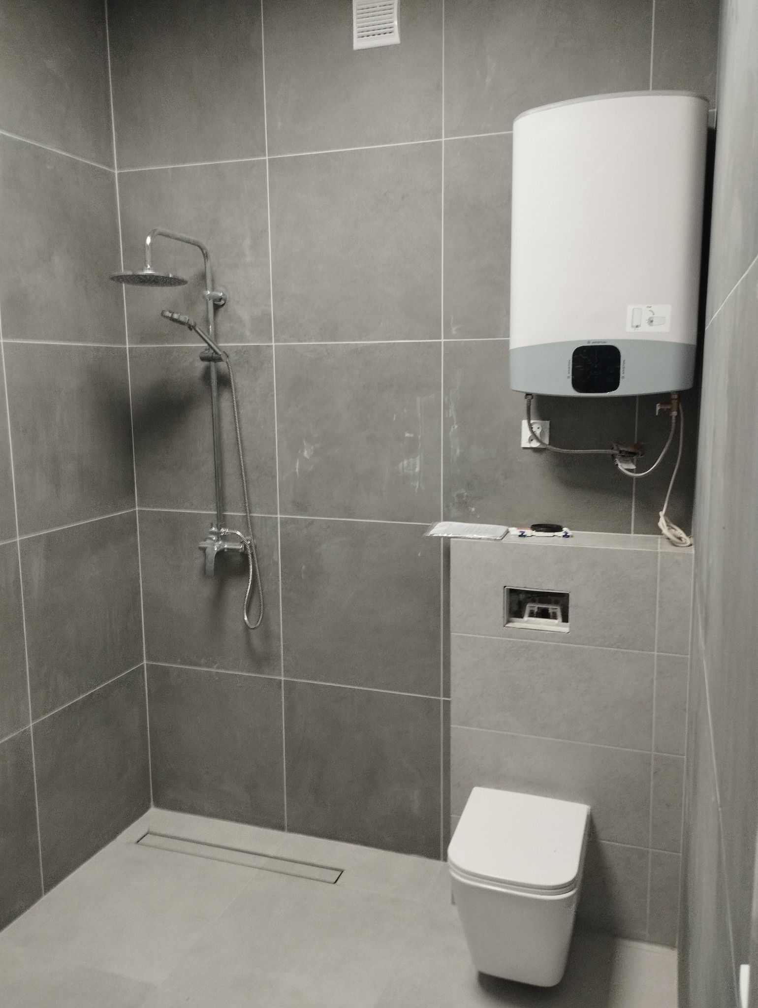 Szara łazienka z kabiną prysznicową typu walk-in, podwieszaną toaletą i bojlerem elektrycznym, wykończona płytkami w jednolitym odcieniu szarości.