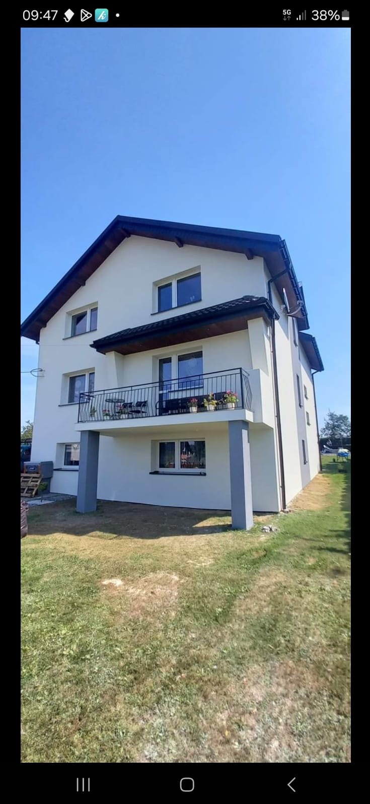 Dwupiętrowy dom z jasną elewacją, czarnym dachem i balkonem z czarną balustradą, otoczony trawnikiem.