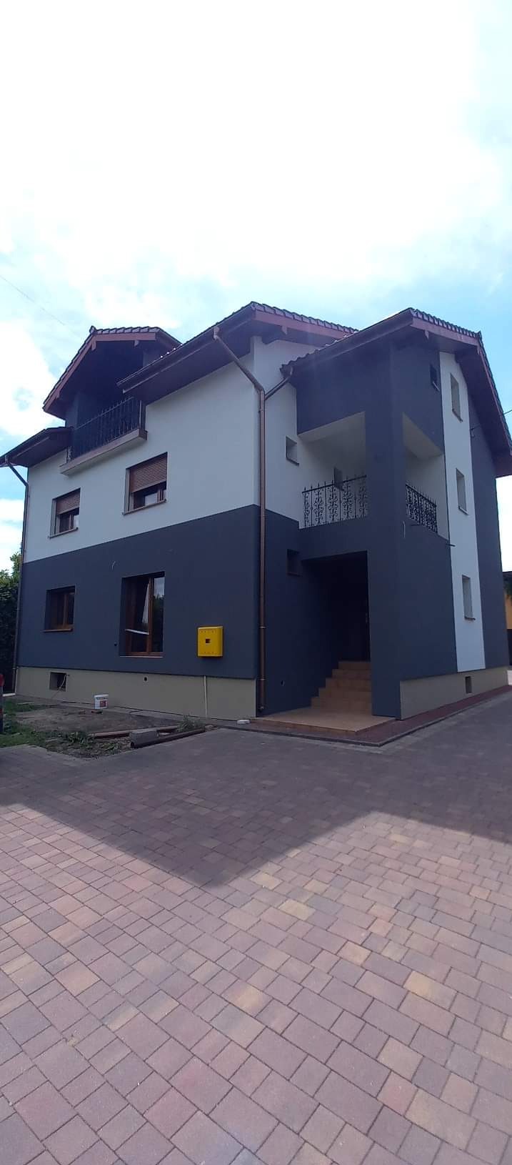 Dwukondygnacyjny dom z elewacją w dwóch odcieniach szarości, z ceglaną kostką brukową na podjeździe i żółtą skrzynką pocztową na ścianie.