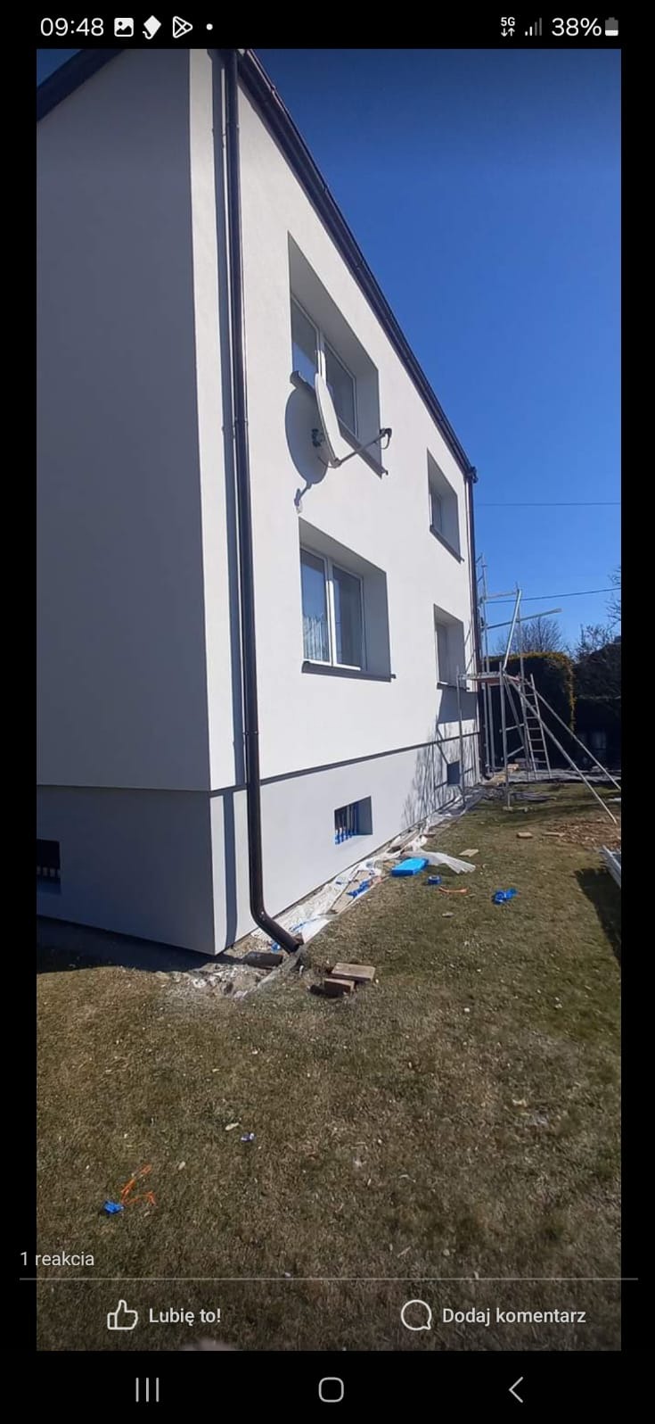 Dwupiętrowy dom z białą elewacją, brązowymi rynnami i anteną satelitarną, widok z boku na tle błękitnego nieba, z widocznymi rusztowaniami i przygotowanym terenem wokół fundamentów.