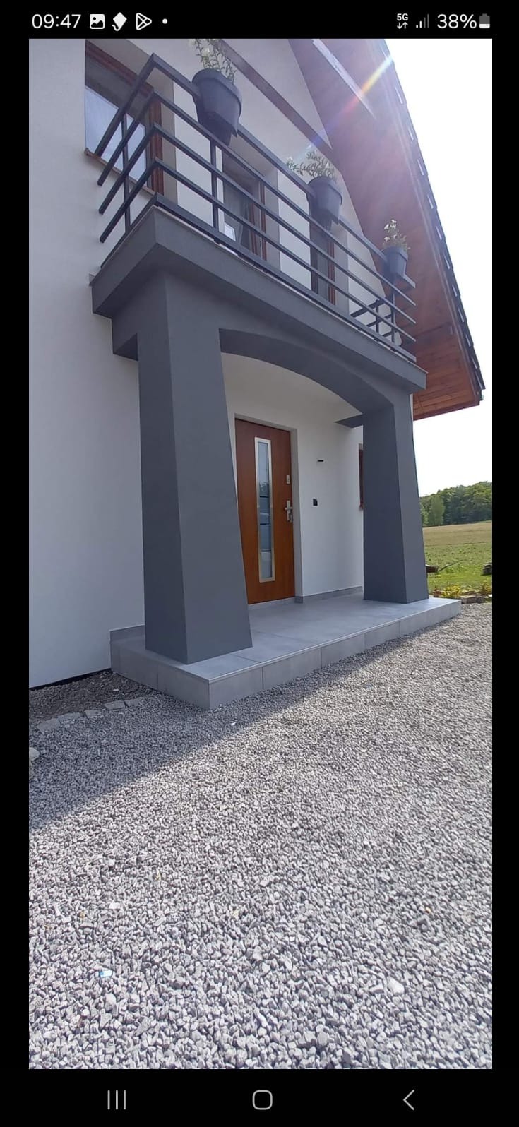 Wejście do domu jednorodzinnego w Jastrzębiu-Zdroju z zadaszeniem wspartym na kolumnach, balkonem na piętrze z czarną balustradą i donicami z kwiatami na nim. Podłoże wyłożone drobnym żwirem.