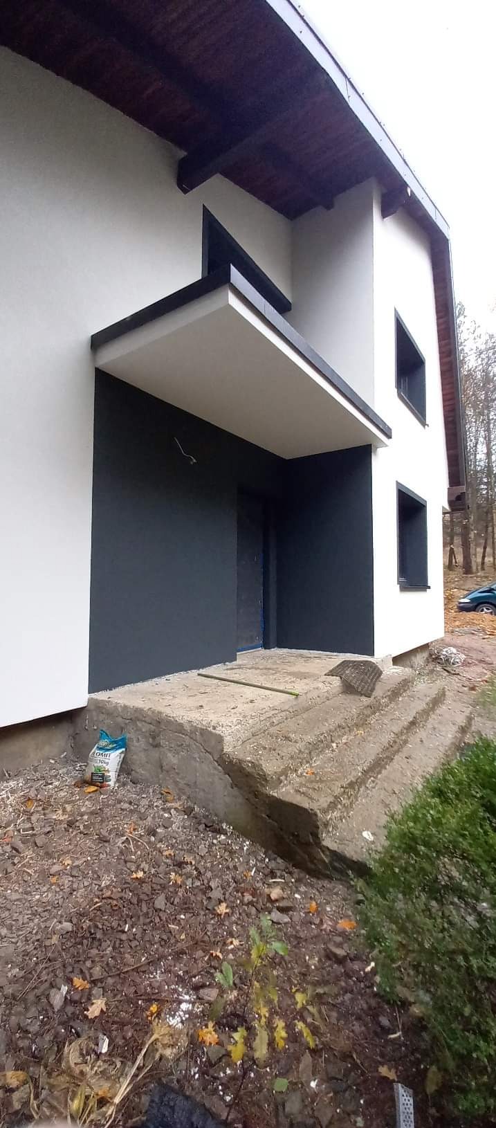 Dwukolorowa elewacja domu jednorodzinnego z betonowymi schodami wejściowymi, widoczne ślady prac budowlanych i worek zaprawy.