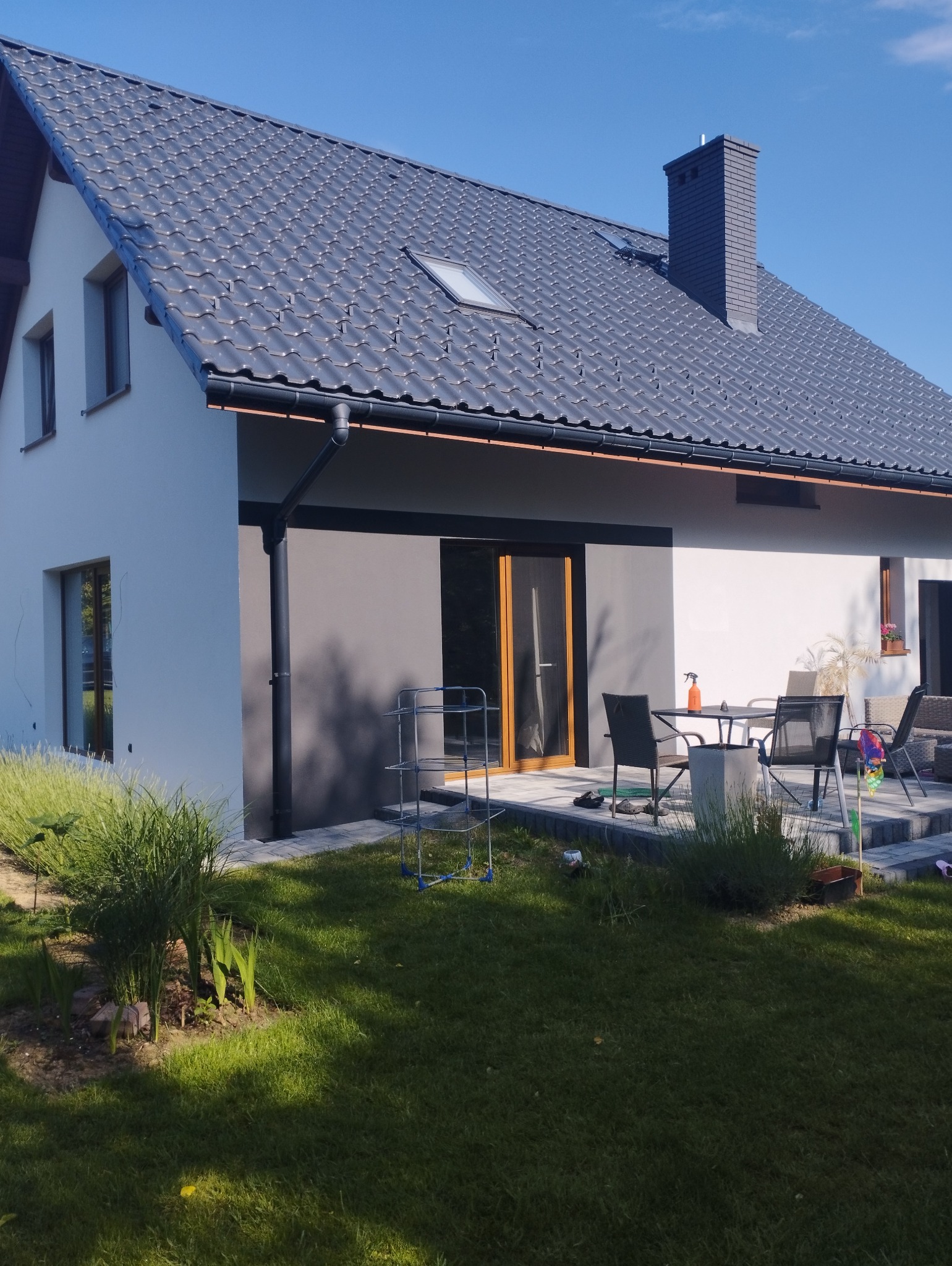 Nowoczesny dom jednorodzinny z dwukolorową elewacją (białą i szarą), grafitowy dach z lukarną, taras z meblami ogrodowymi i zielony trawnik.