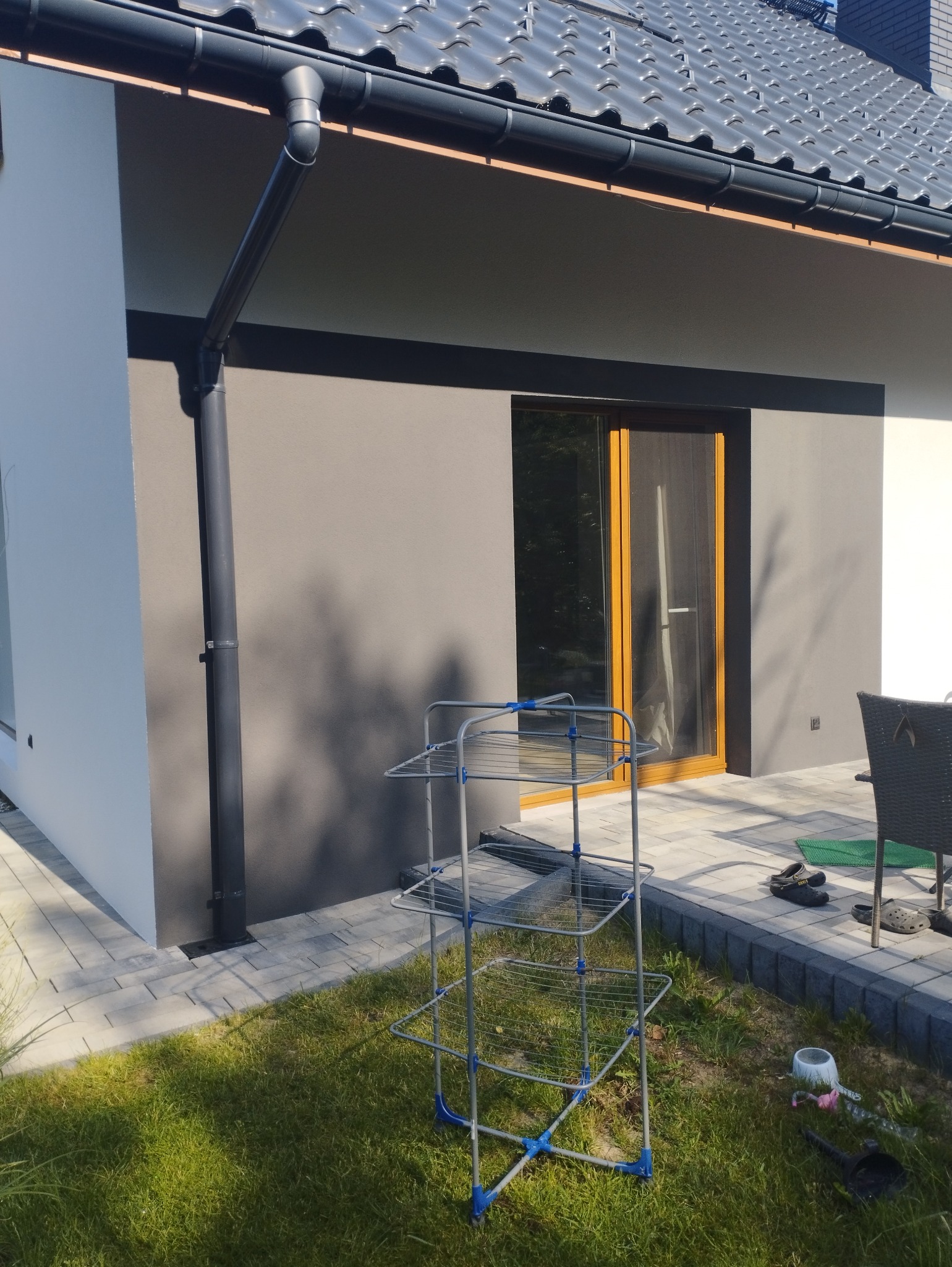 Nowoczesny dom z szarą elewacją, czarną stolarką okienną i rynną, widok na fragment elewacji i otoczenie z trawnikiem, na pierwszym planie suszarka na pranie.