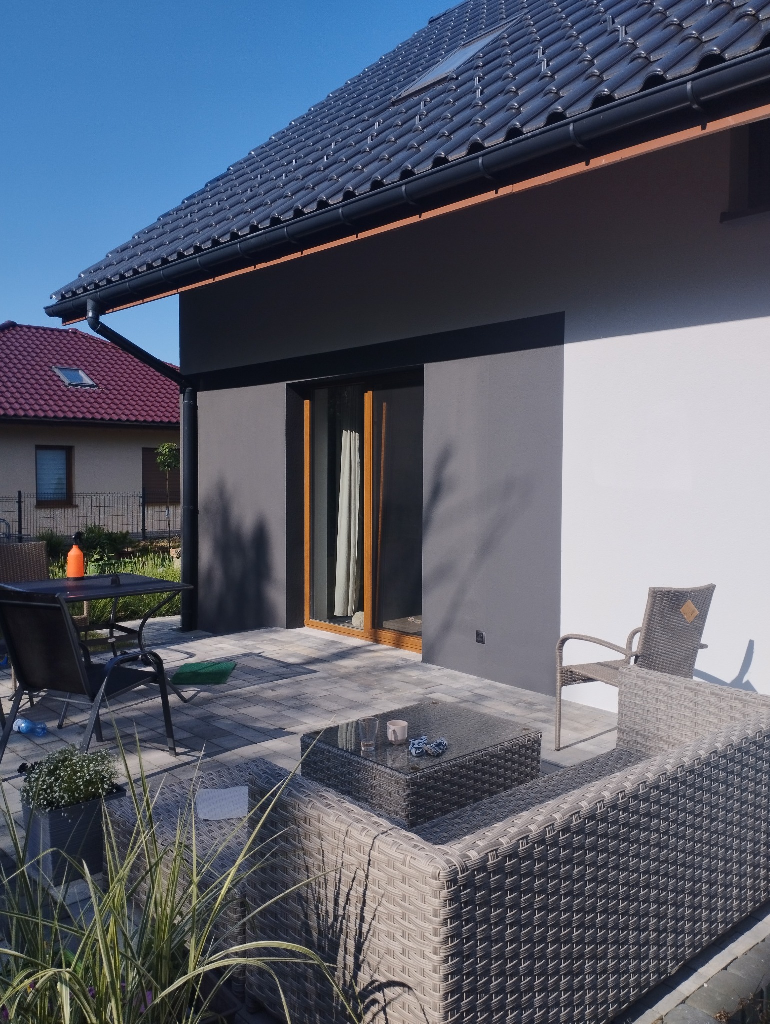Nowoczesny dom z grafitowo-białą elewacją, ciemną dachówką i tarasem z meblami ogrodowymi w słoneczny dzień.