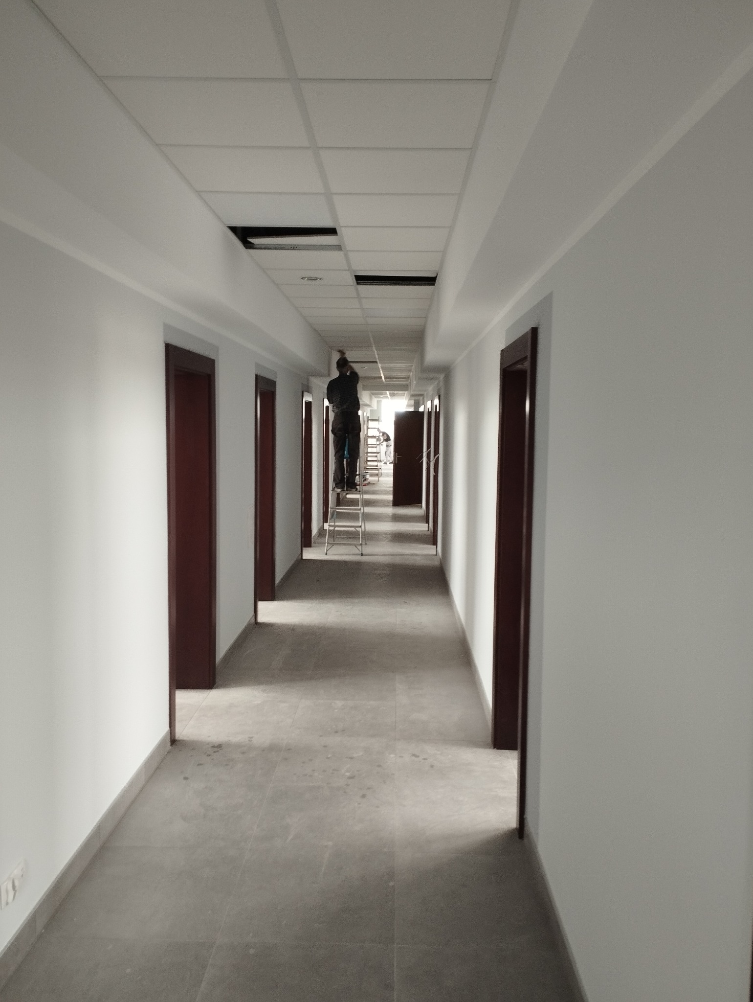 Pracownik na drabinie maluje sufit w długim korytarzu z białymi ścianami i brązowymi drzwiami, widoczne niedokończone fragmenty sufitu.