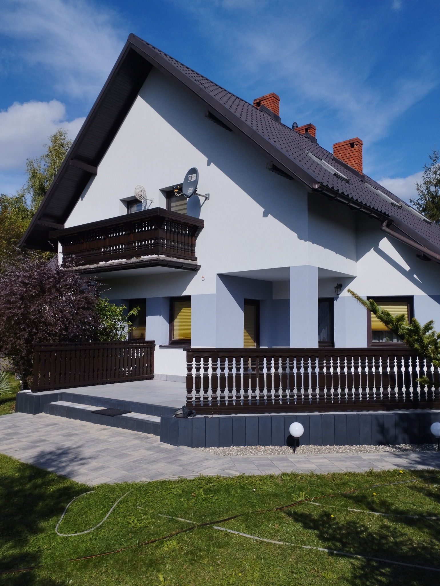 Dwukondygnacyjny dom z biało-szarą elewacją, brązowym dachem, balkonem z drewnianą balustradą i zadbanym trawnikiem z kostką brukową.
