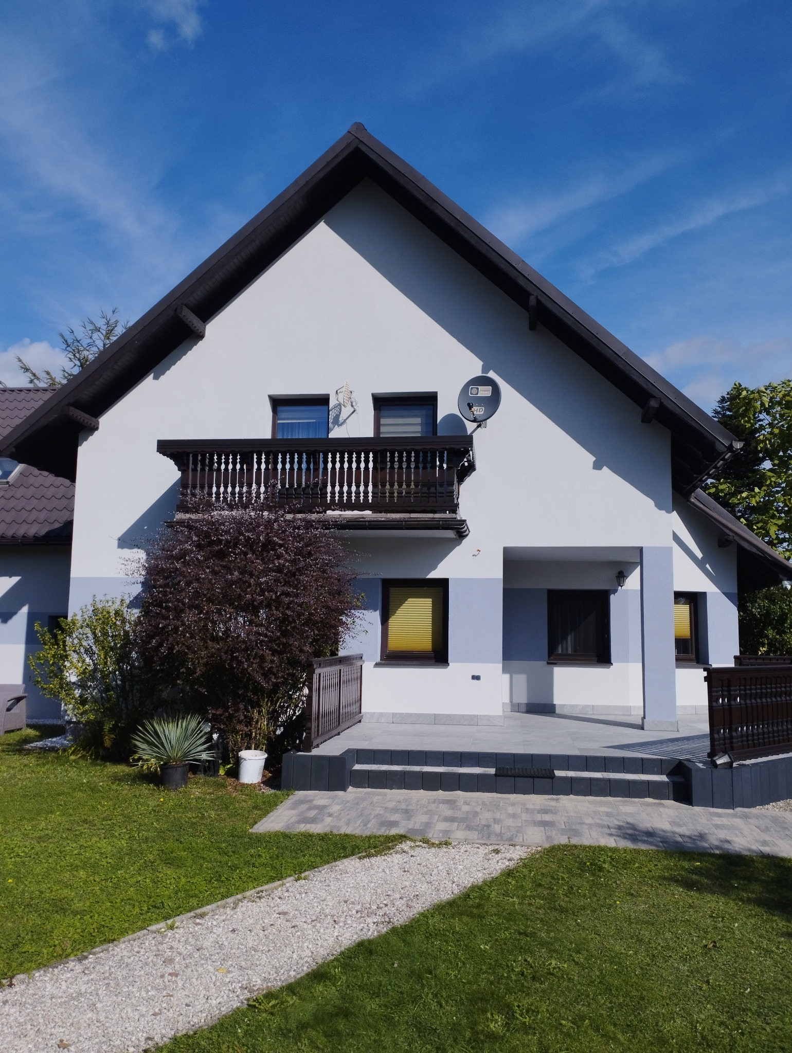 Dwukondygnacyjny dom jednorodzinny z balkonem z drewnianą balustradą, białą elewacją z szarymi akcentami, brązowym dachem i anteną satelitarną na tle błękitnego nieba. Wokół domu zadbany trawnik...