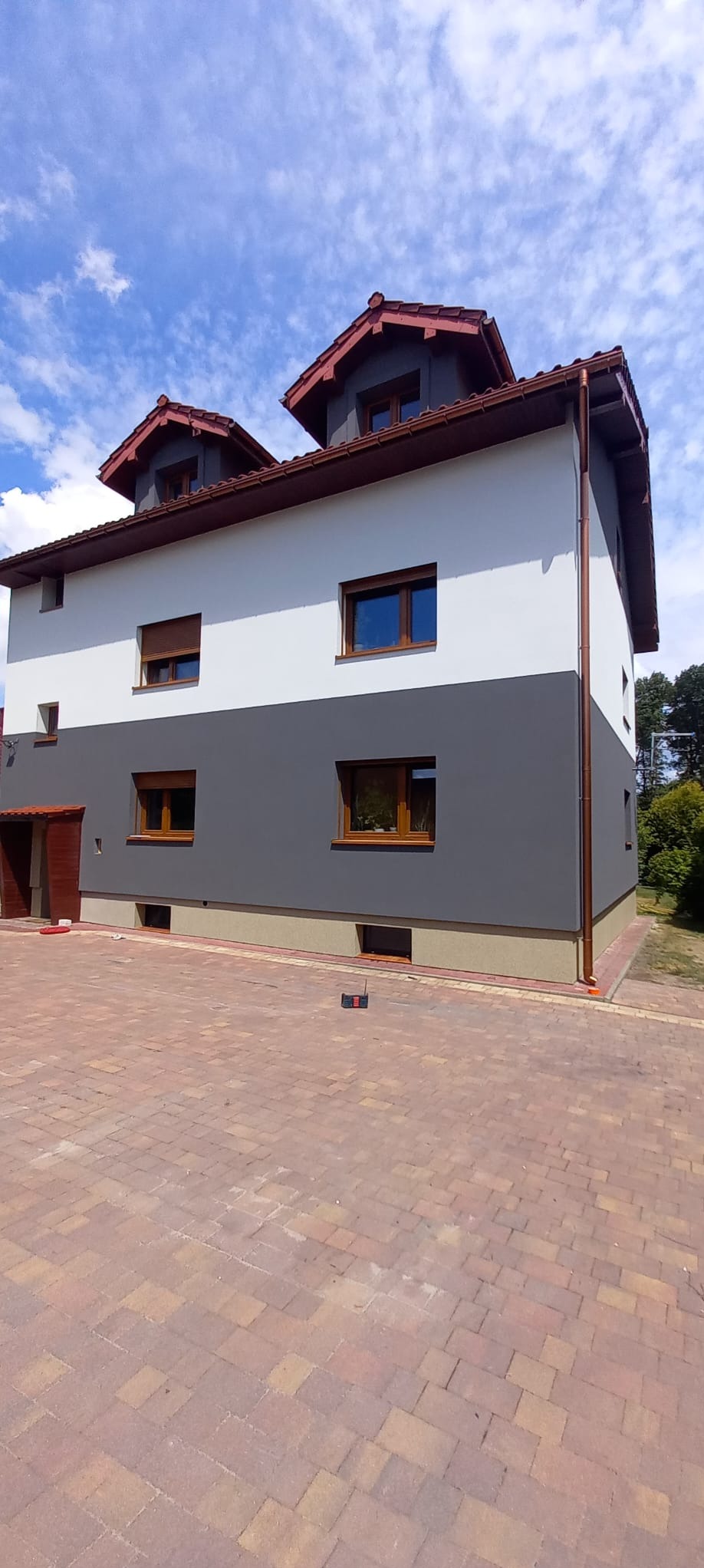 Dwukondygnacyjny dom z elewacją w dwóch odcieniach szarości, z brązowymi oknami i dachem, widziany z poziomu brukowanego podjazdu w słoneczny dzień.