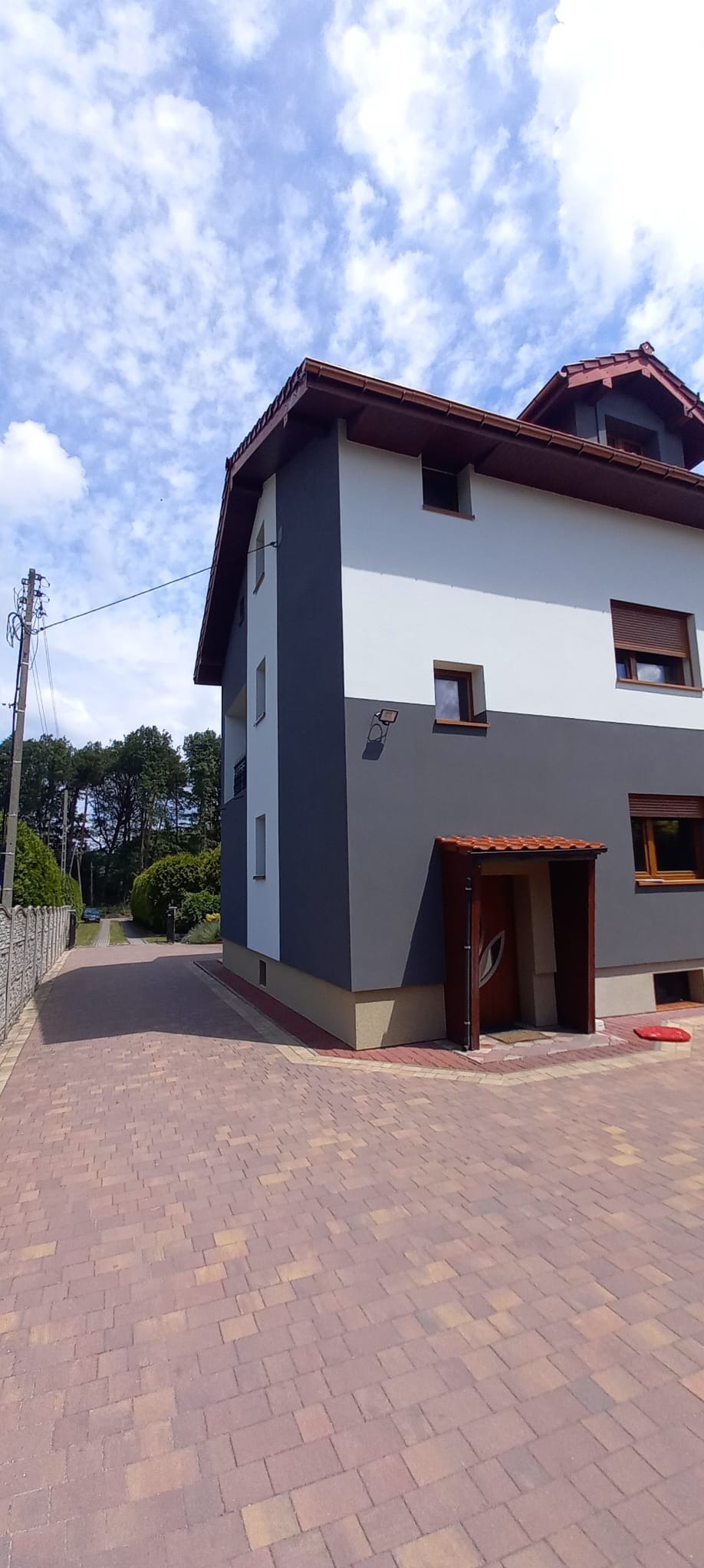 Dwukondygnacyjny dom z elewacją w odcieniach szarości i bieli, z brązowymi oknami i zadaszeniem nad wejściem, sfotografowany z poziomu brukowanego podjazdu w słoneczny dzień.