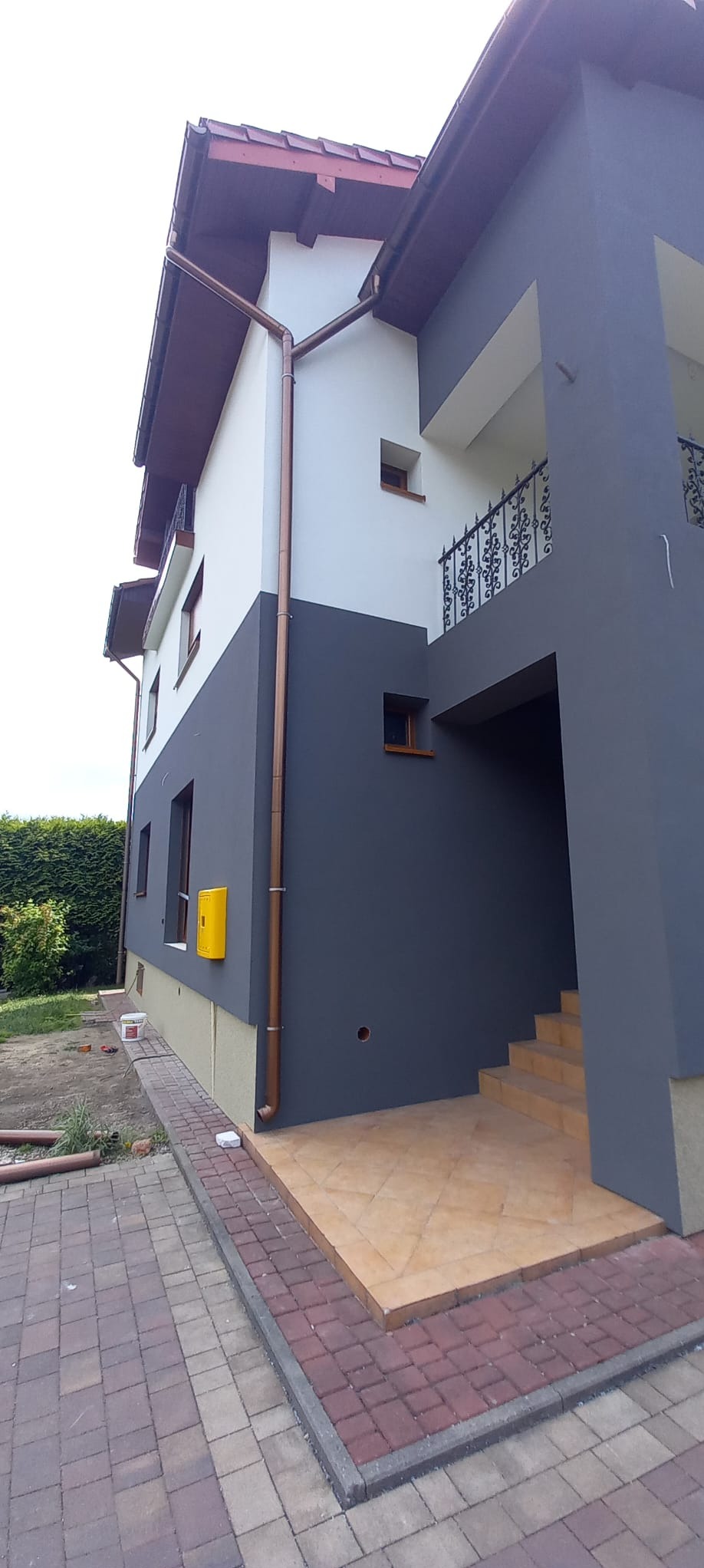 Dwukolorowa elewacja domu z żółtą skrzynką gazową, widok z poziomu chodnika z kostki brukowej.