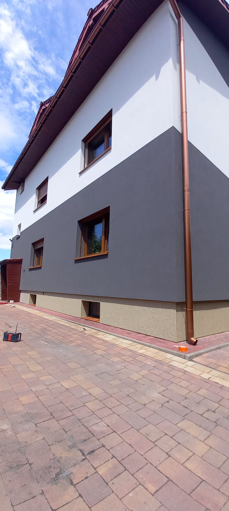 Dwukondygnacyjny dom z elewacją w trzech odcieniach: biały na górze, szary pośrodku i beżowy na dole, z brązowymi oknami i rynnami. Widoczny fragment chodnika z kostki brukowej.