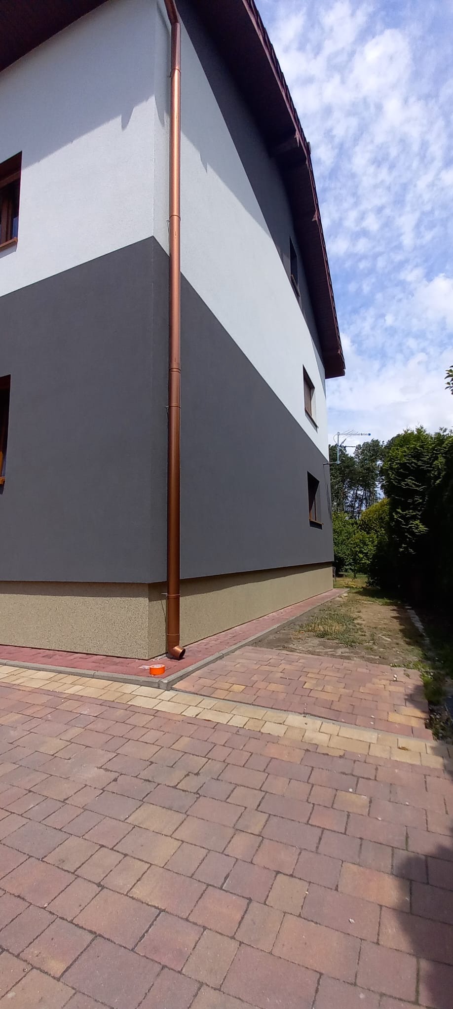 Dwukolorowa elewacja domu z brązową rynną, widok z boku na narożnik budynku, kostka brukowa na podwórku.