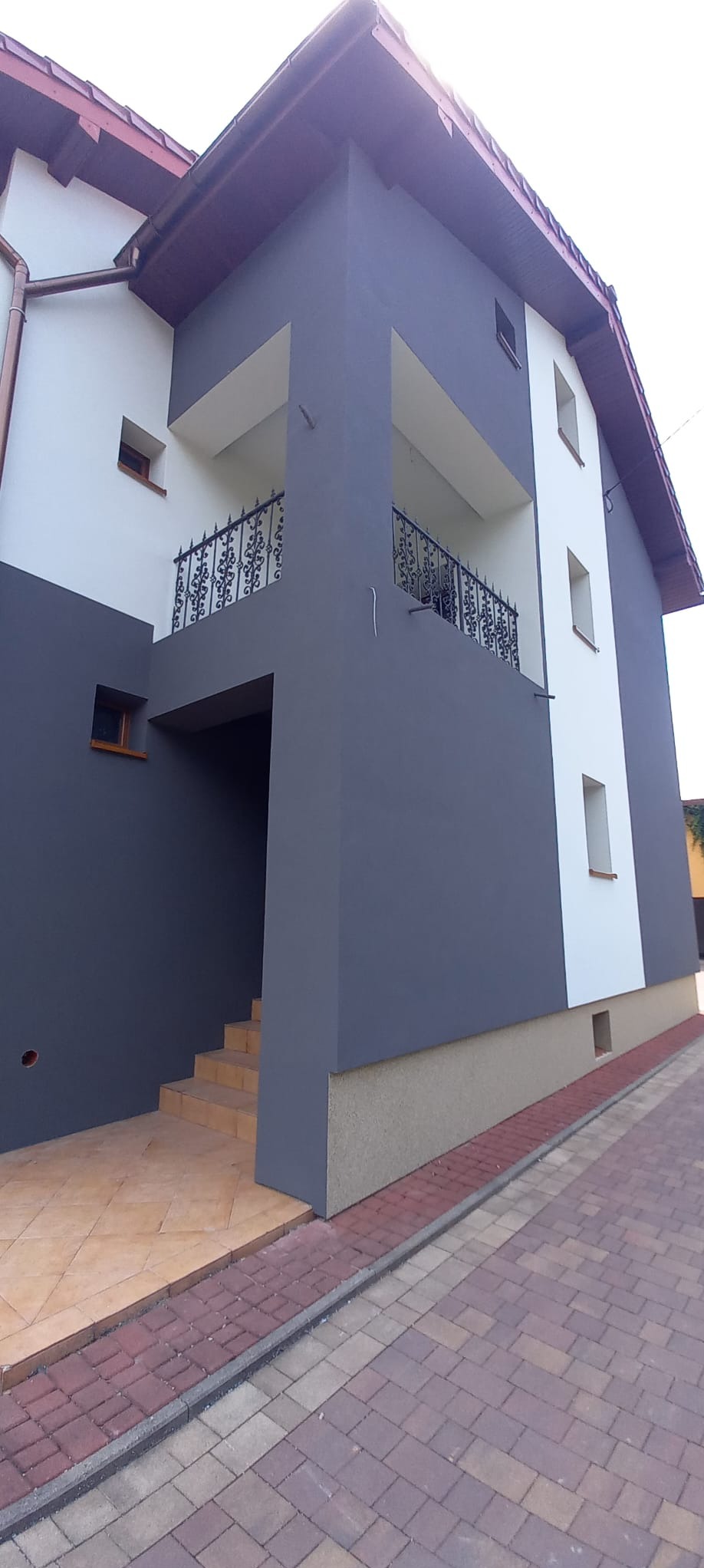 Dwukondygnacyjny dom z szarą i białą elewacją, balkon z ozdobną, metalową balustradą i schody wejściowe z beżowymi płytkami.