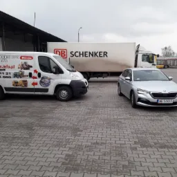 Flota pojazdów na szarym brukowanym placu: biała furgonetka z grafiką systemów monitoringu GPS i kontroli paliwa, ciężarówka DB Schenker i srebrna Skoda z tablicą rejestracyjną WE 544XV.