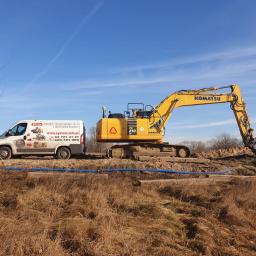 P.P.H.U. SYSTEM - Żółta koparka Komatsu PC210LC w trakcie prac ziemnych, obok biały bus z reklamą systemów monitoringu GPS i kontroli paliwa, błękitne rury na ziemi.