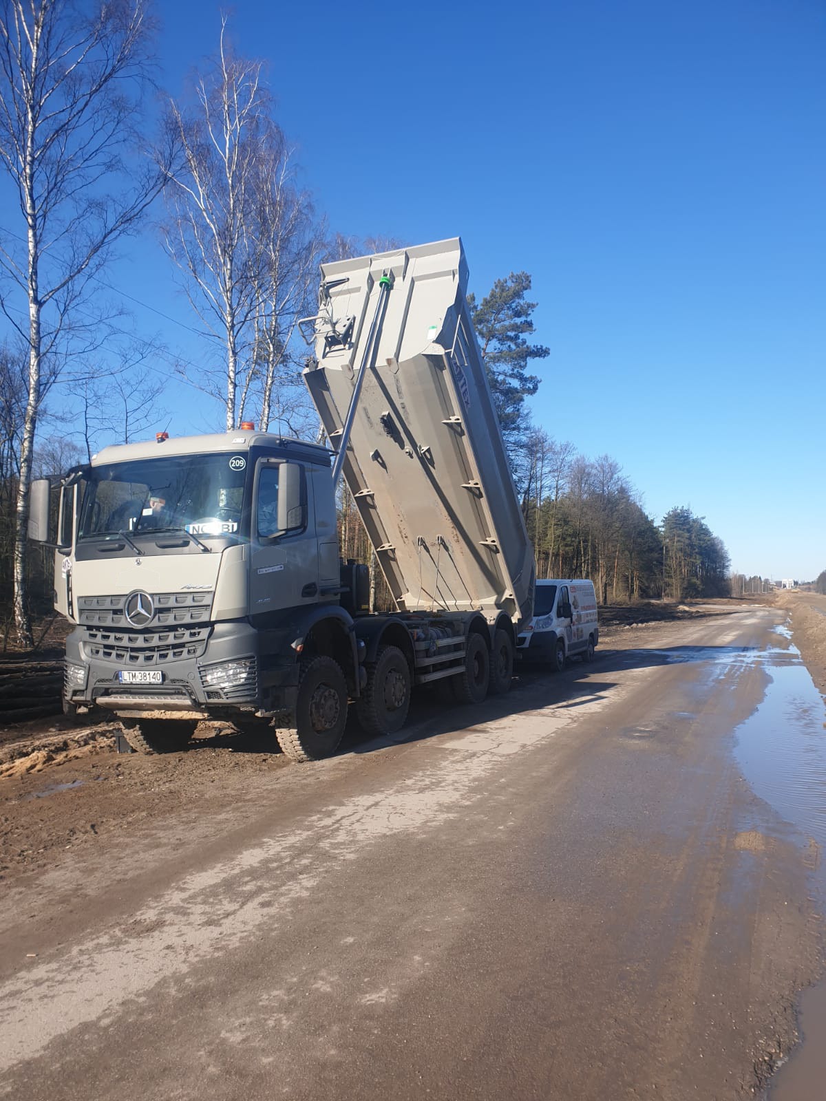 Ciężarówka Mercedes-Benz z podniesioną naczepą na błotnistej drodze, obok zaparkowany biały bus, w tle drzewa i błękitne niebo.