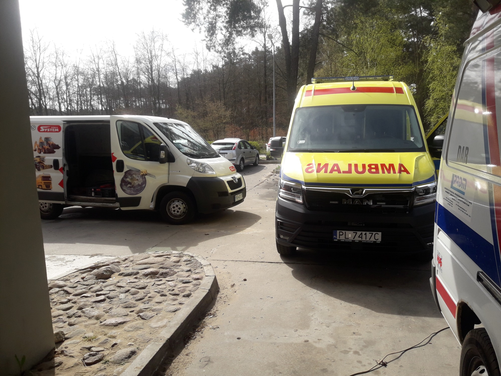Dwa ambulanse i biała furgonetka z otwartymi drzwiami na parkingu, w tle drzewa bez liści.