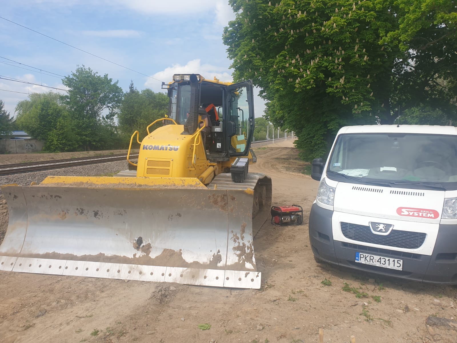 Żółty spychacz Komatsu z brudnym lemieszem stoi obok białego busa z logo firmy System na tle torów kolejowych i drzew