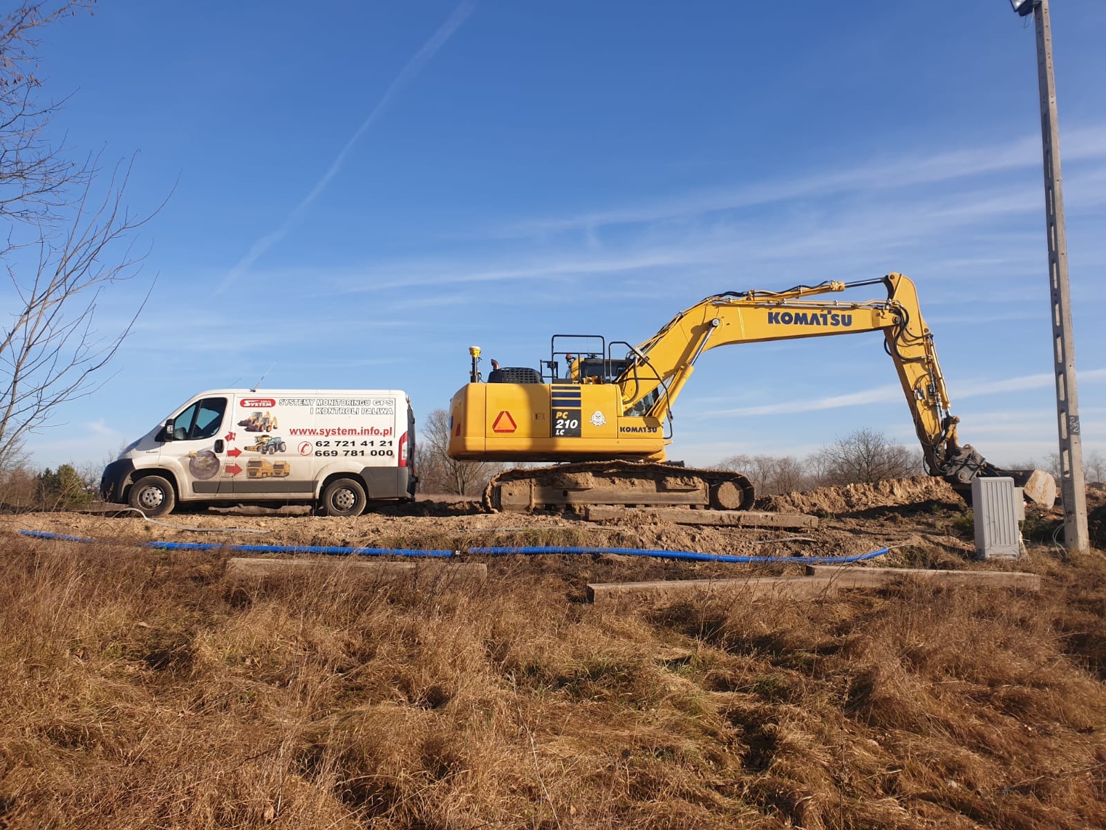 Żółta koparka Komatsu PC210LC w trakcie prac ziemnych, obok biały bus z reklamą systemów monitoringu GPS i kontroli paliwa, błękitne rury na ziemi.