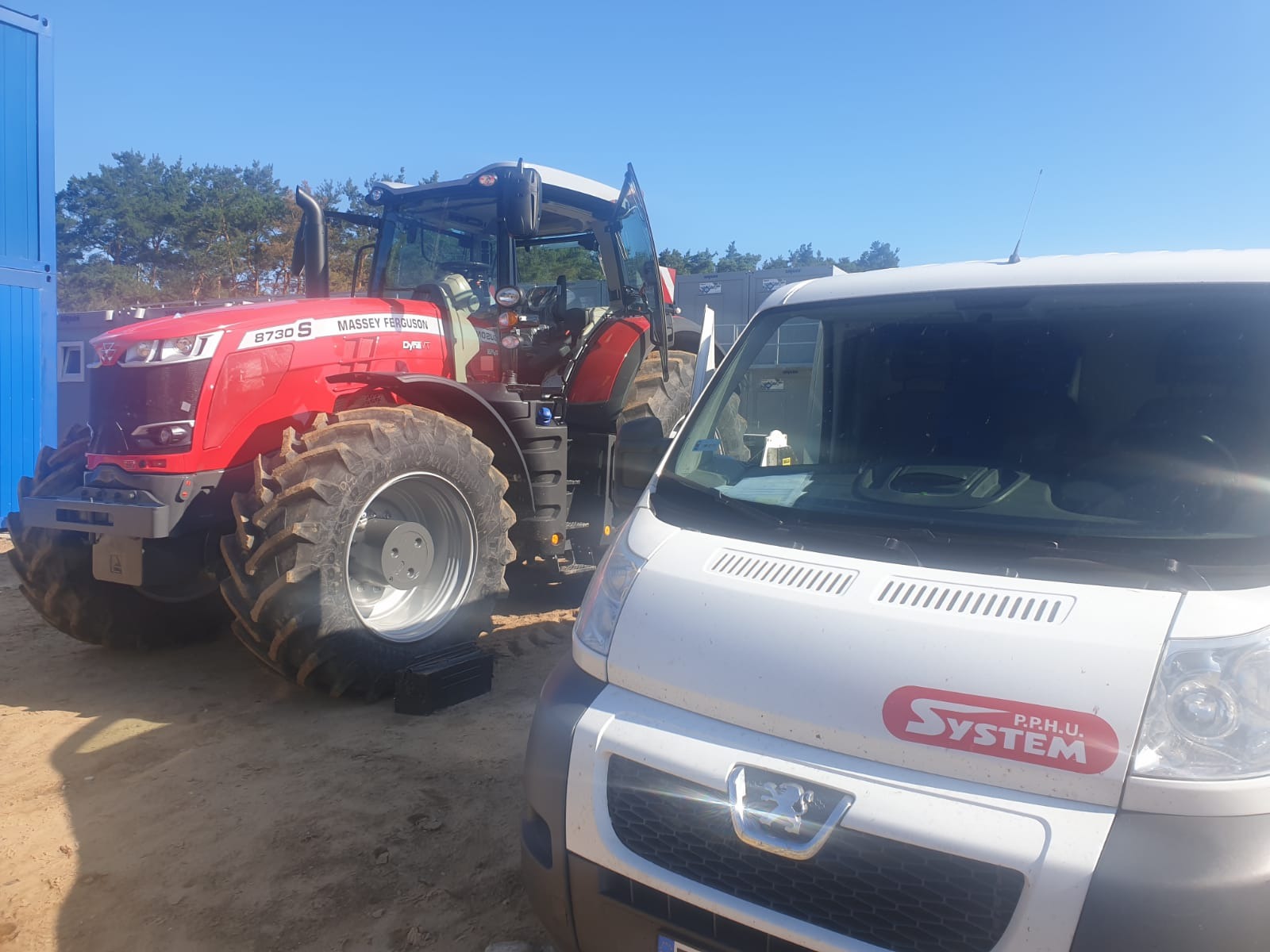 Czerwony ciągnik rolniczy Massey Ferguson 8730S z otwartymi drzwiami kabiny, zaparkowany obok białego samochodu dostawczego z logo firmy SYSTEM P.P.H.U. na tle piaszczystego terenu i błękitnego nieba.