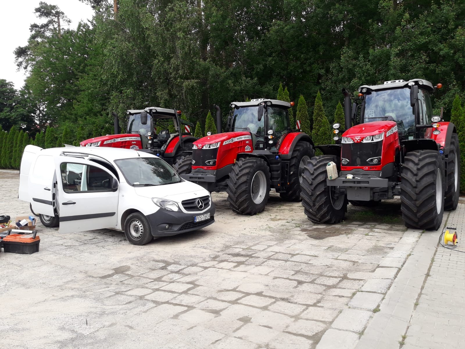 Trzy czerwone ciągniki Massey Ferguson na betonowym placu, biały samochód serwisowy Mercedes z otwartymi drzwiami i skrzynką narzędziową obok.