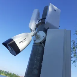 Kamera monitoringu Hikvision HiWatch Series zamontowana na metalowym słupie z antenami, widok z dołu na tle błękitnego nieba i jeziora.