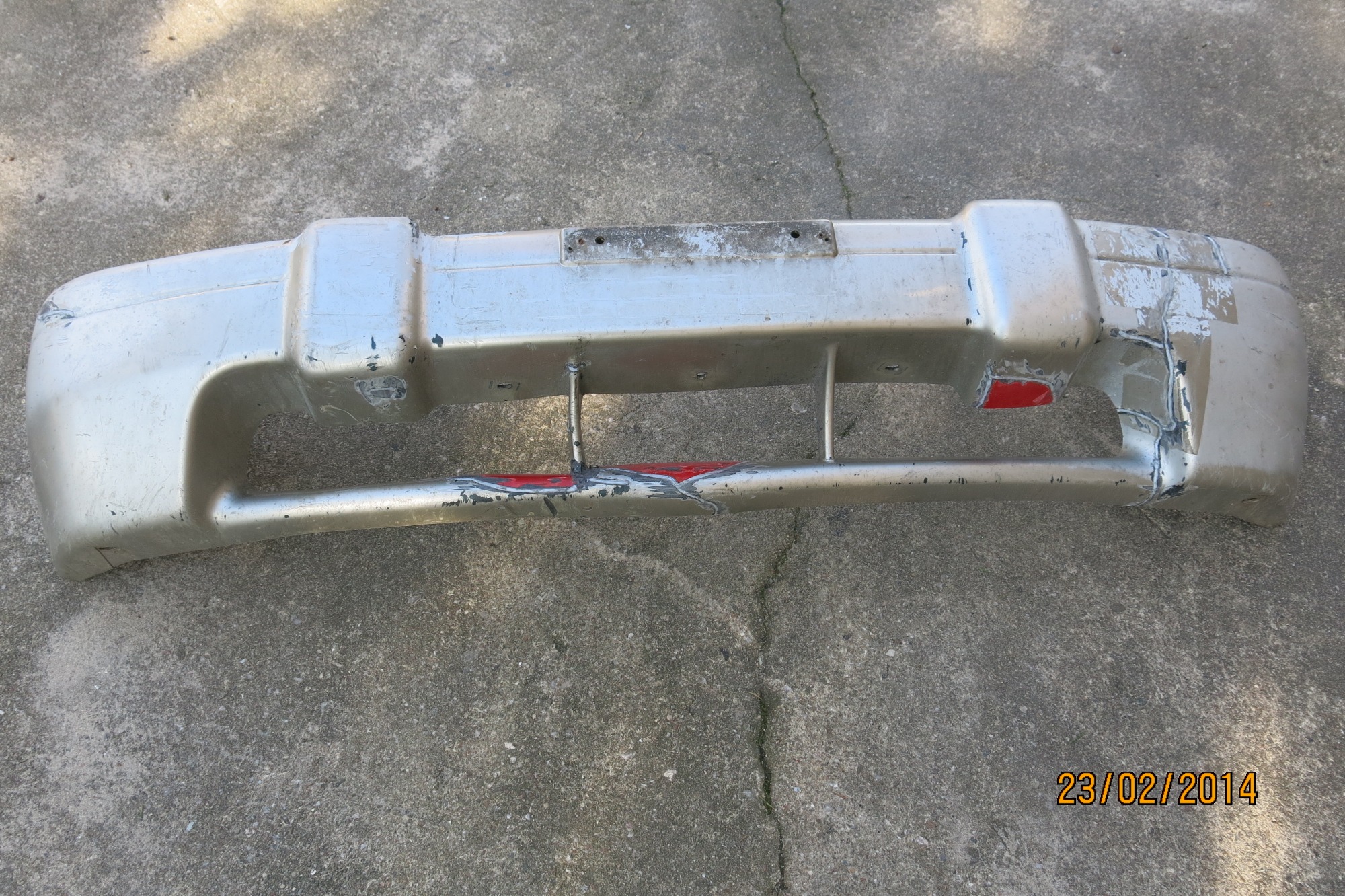 Zdemontowany, uszkodzony, srebrny zderzak samochodowy z widocznymi śladami naprawy i rdzawymi plamami, leżący na betonowym podłożu.