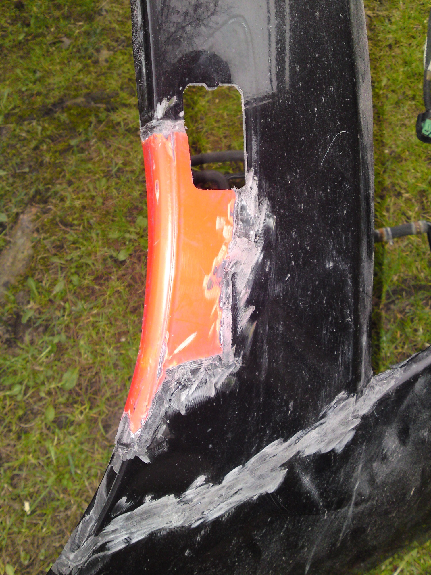 Naprawa pękniętego, czarnego elementu karoserii z widocznym pomarańczowym wzmocnieniem i śladami szpachli na trawiastym tle.