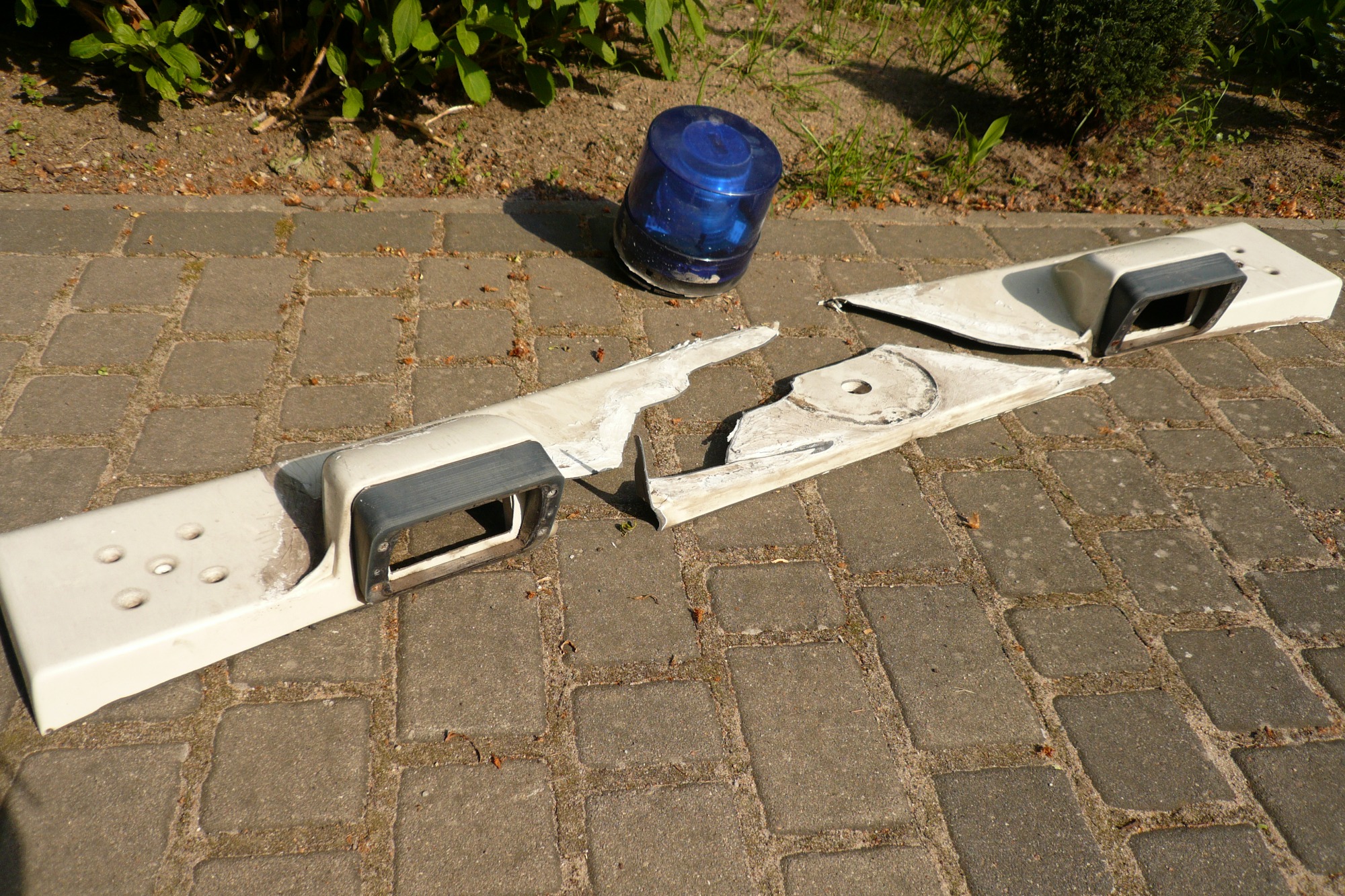 Uszkodzony, biały element karoserii samochodu z niebieskim, okrągłym światłem leżący na szarej kostce brukowej.