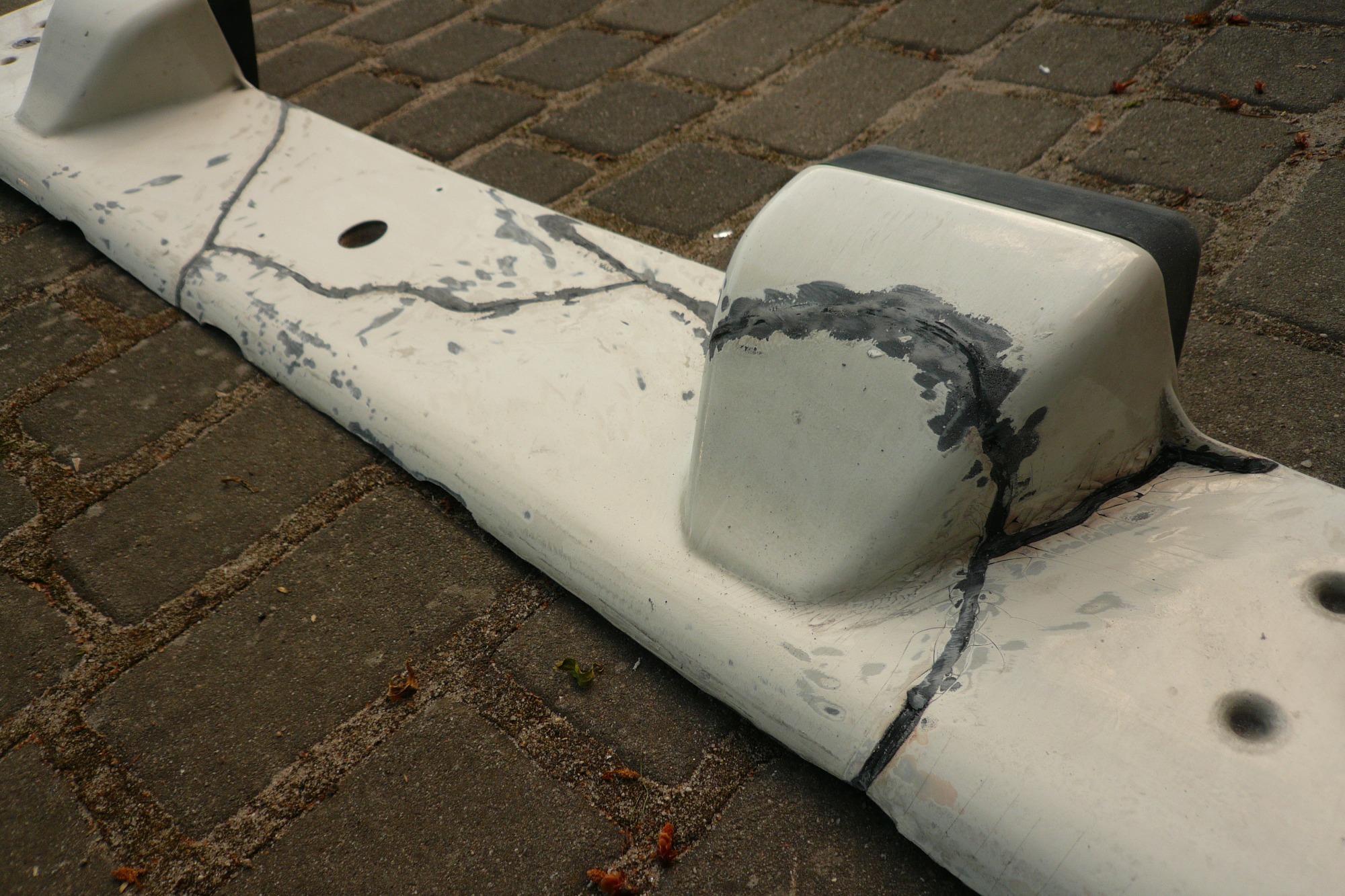 Biały element konstrukcyjny z widocznymi pęknięciami i śladami naprawy wypełnionymi szpachlą, leżący na szarej kostce brukowej.