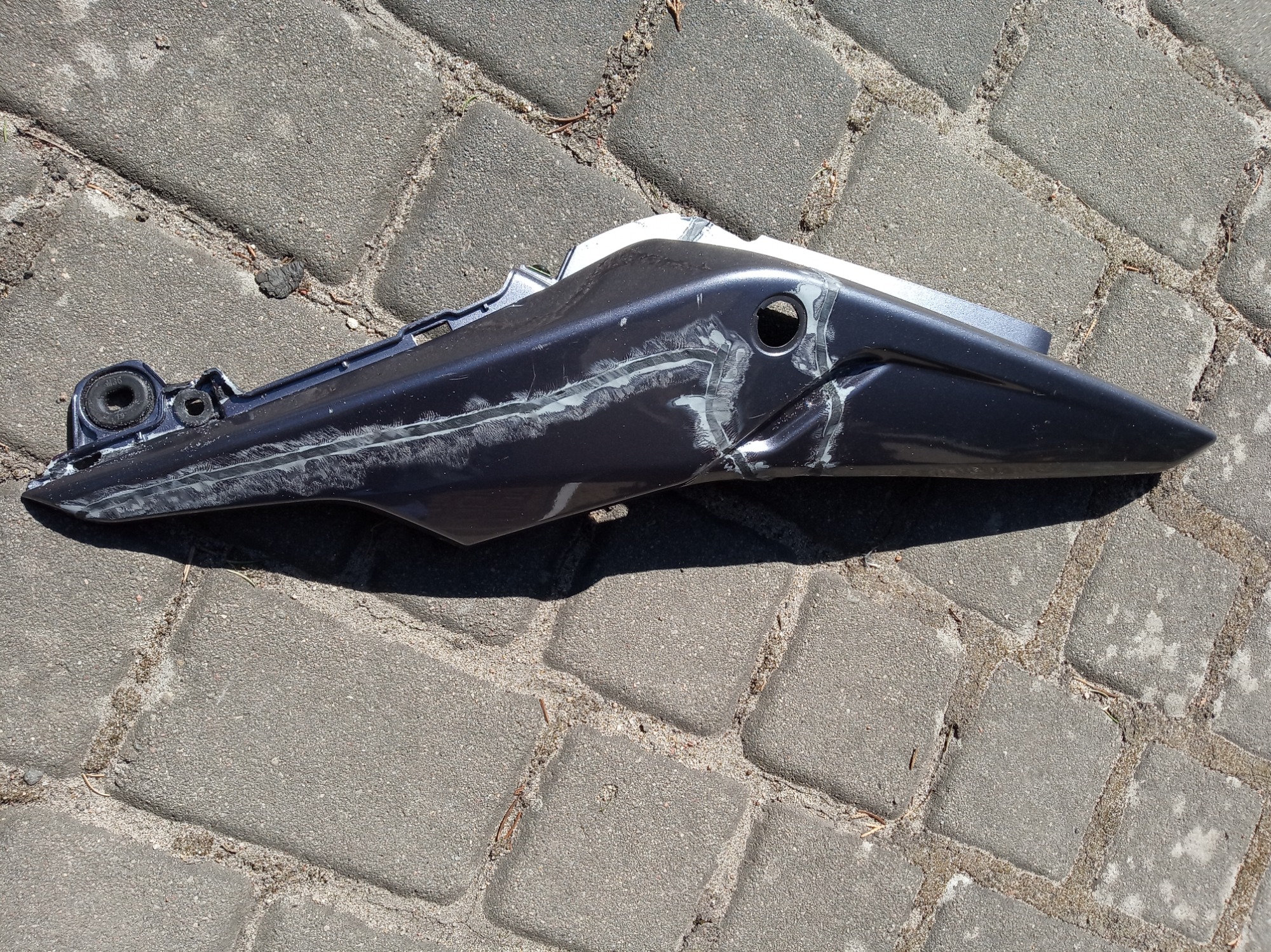 Uszkodzony, szpachlowany element karoserii motocykla w kolorze grafitowym leżący na szarej kostce brukowej.