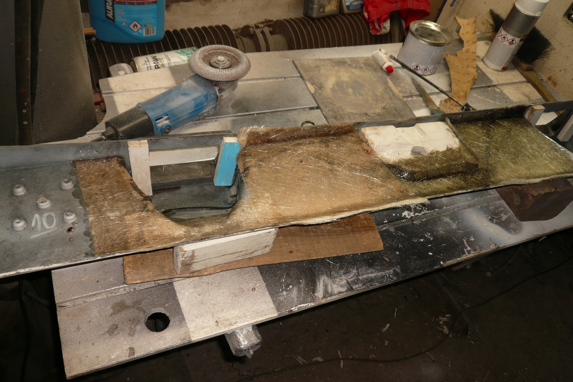 Fragment elementu z włókna szklanego na metalowym stole warsztatowym, widoczne narzędzia i materiały, w tym szlifierka kątowa i puszki z farbą.