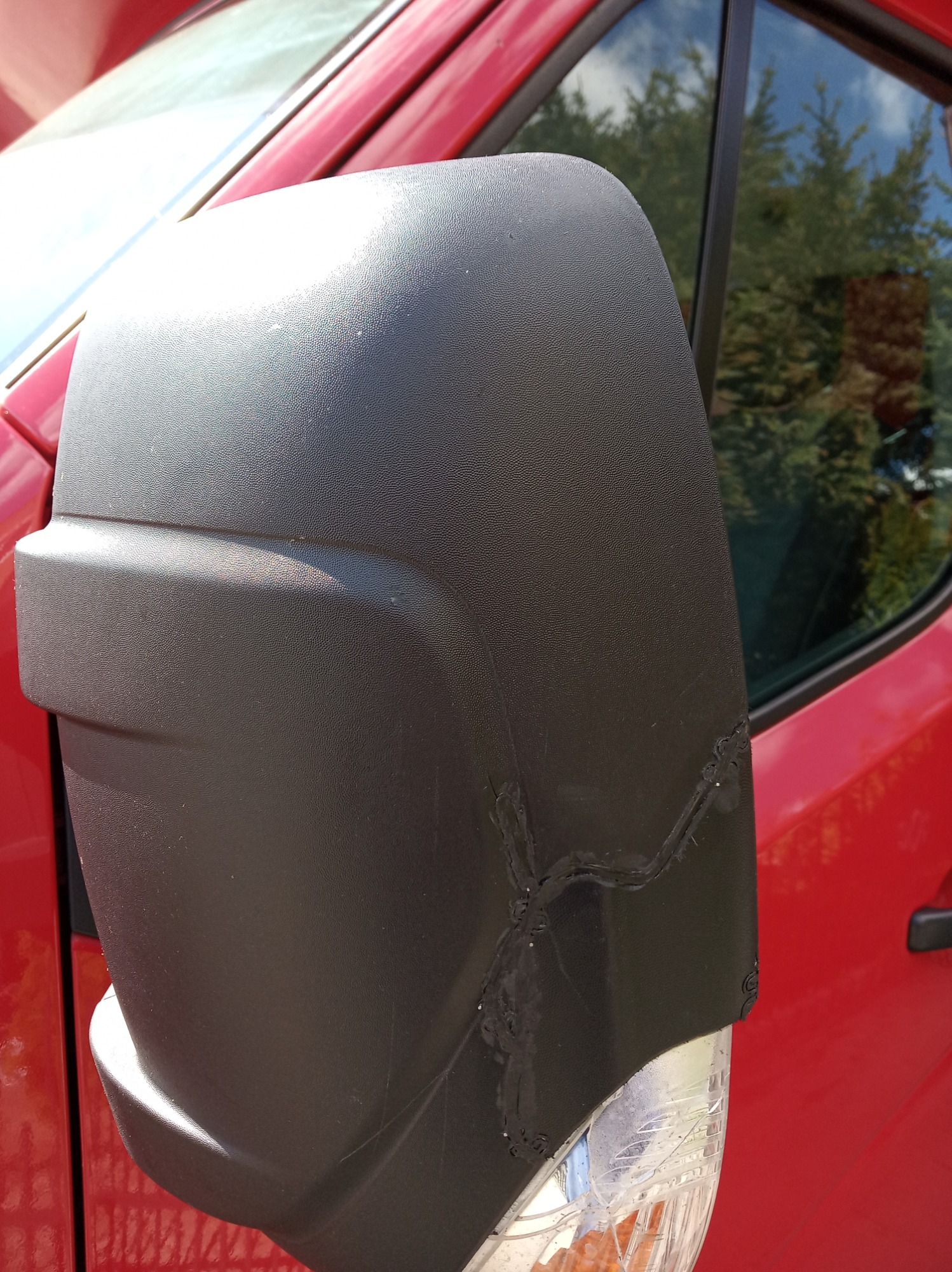 Uszkodzone czarne lusterko boczne czerwonego samochodu, widoczne pęknięcia i ślady naprawy.