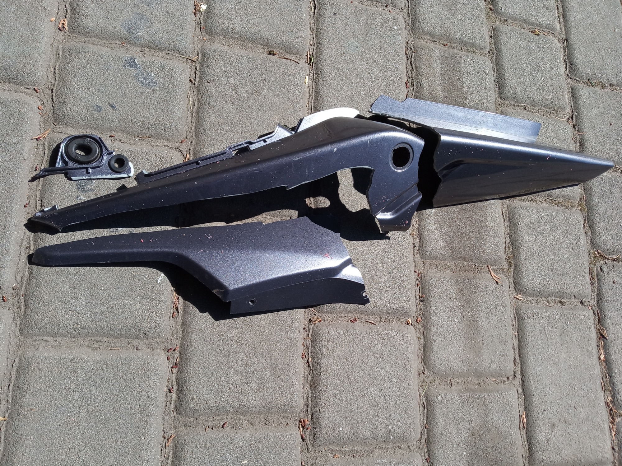 Uszkodzone, plastikowe elementy karoserii motocykla w odcieniach szarości, leżące na szarej kostce brukowej.