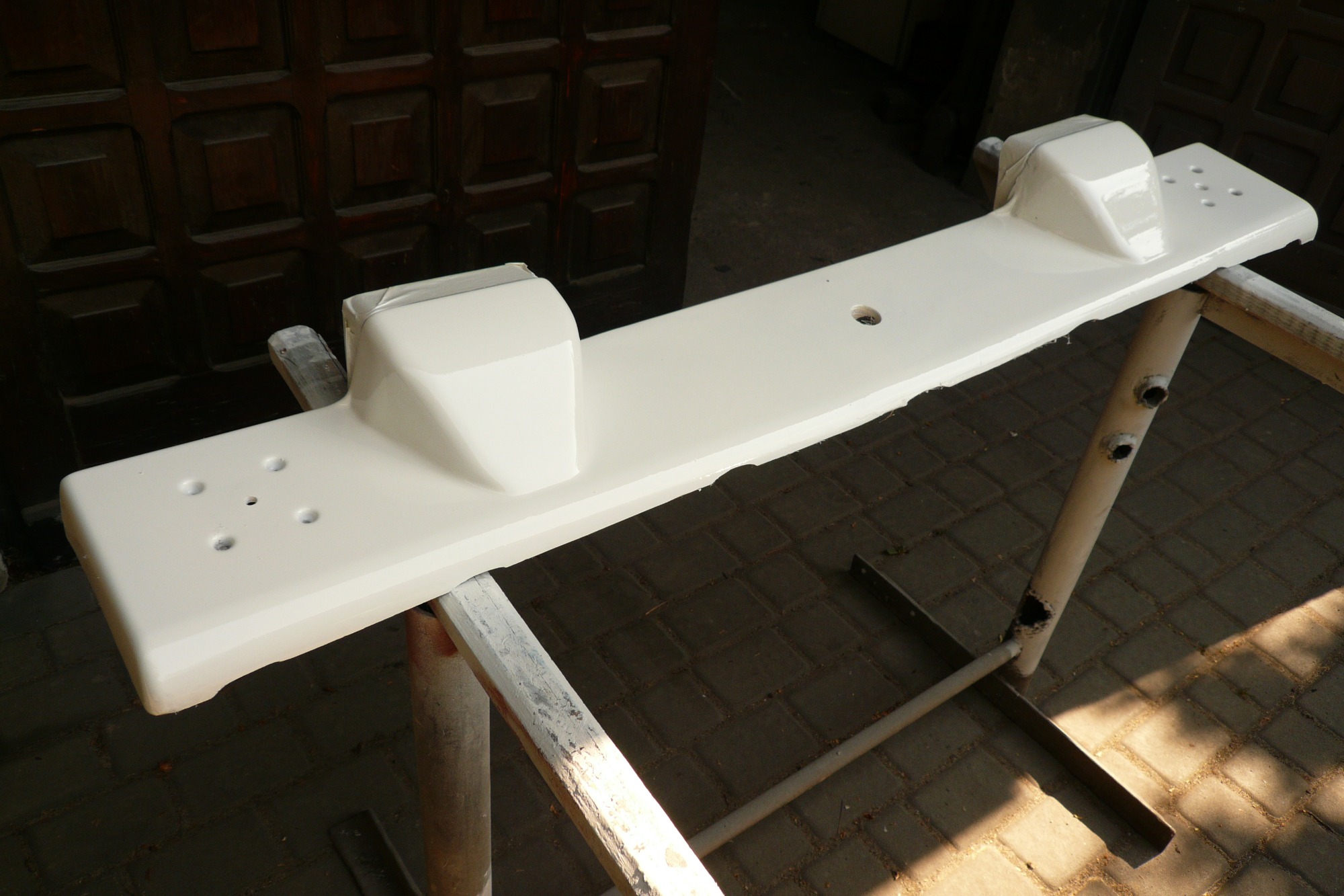 Biały element konstrukcyjny z otworami, prawdopodobnie część łodzi, umieszczony na metalowym stojaku w trakcie malowania lub po nim, z widocznym zacienionym tłem z ciemnego drewna i kostki brukowej.