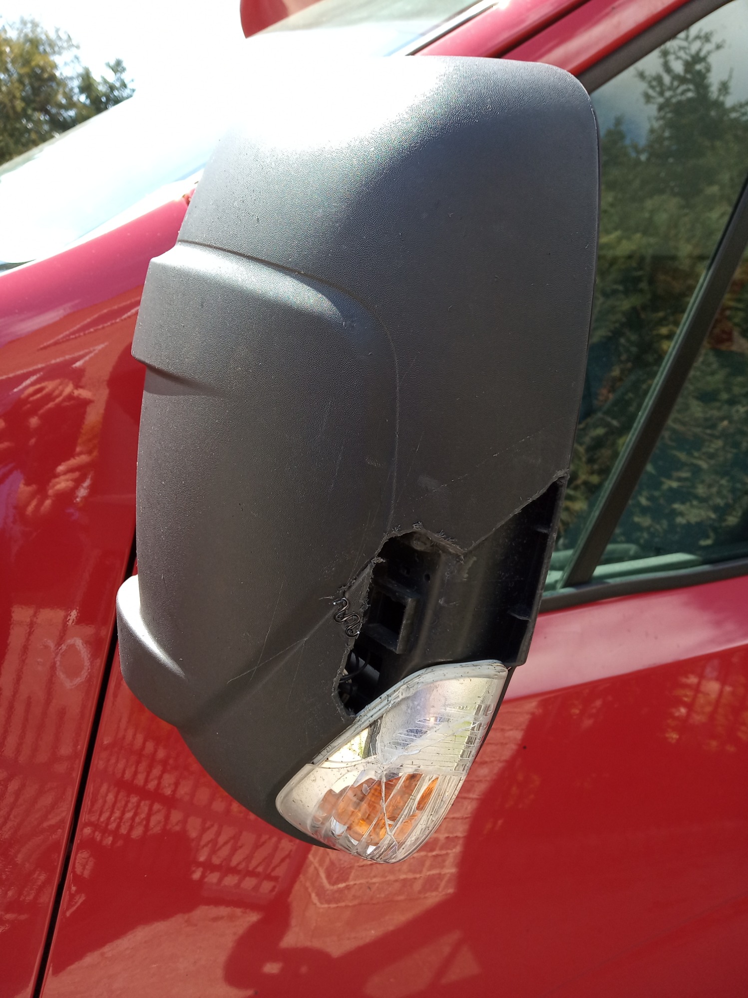 Uszkodzone lusterko boczne czerwonego samochodu w Trzciance, z widocznym pęknięciem obudowy i zarysowaniami.