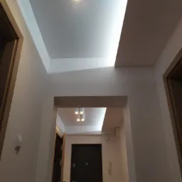 Korytarz z białymi ścianami, ukrytym oświetleniem sufitowym i drewnianymi ościeżnicami drzwi, widziany z dołu.