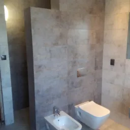 Nowoczesna łazienka z szarymi, wielkoformatowymi płytkami, wiszącą toaletą i bidetem z chromowaną armaturą.